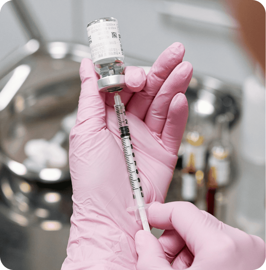 A person in pink gloves, likely a primary care professional, uses a syringe to draw liquid from a vial.