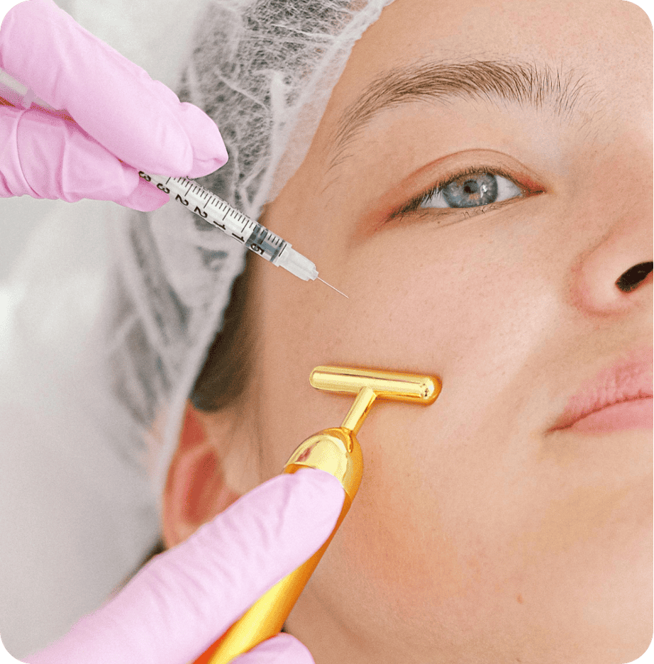 A person receiving a facial injection with a syringe while another person holds a gold facial roller. The patient, wearing a hair cover, is being attended to by the practitioner in pink gloves. This treatment, akin to primary care for the skin, ensures both safety and rejuvenation.