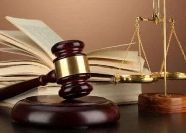 A gavel rests on its base in the foreground, with an opened book and a set of scales in the background, symbolizing the balance and judgment often exercised in primary care decisions.