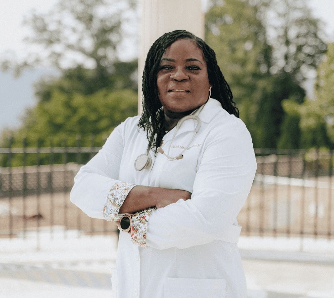 A primary care physician wearing a white lab coat and a stethoscope around her neck stands outdoors with her arms crossed, smiling with trees and a blurred background behind her.