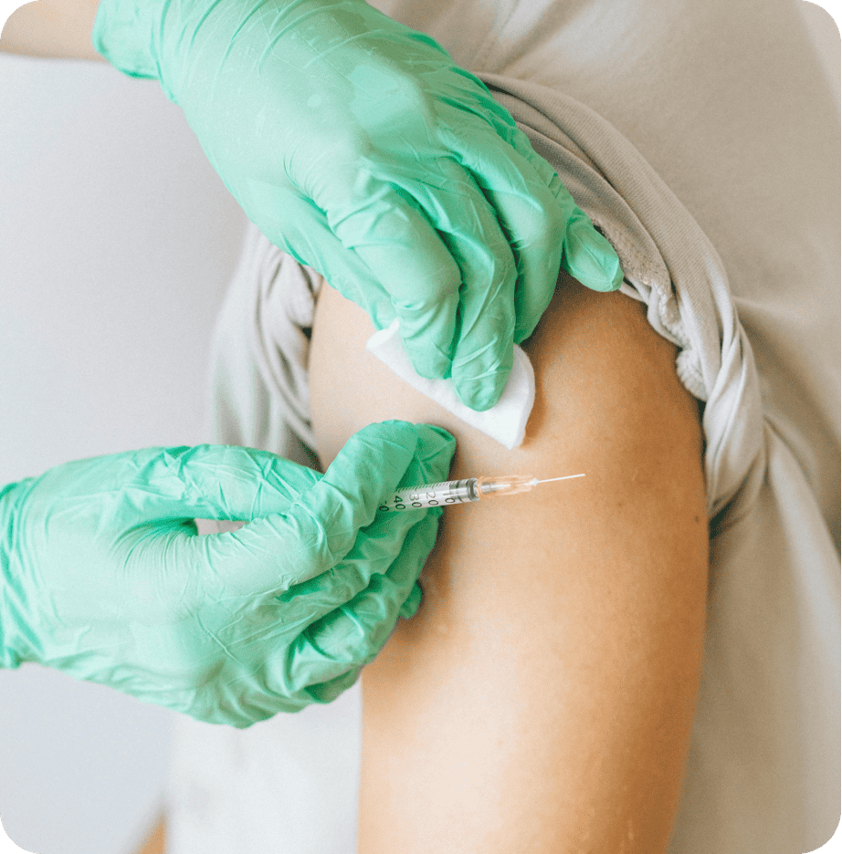 A primary care professional wearing green gloves administers an injection into a person's upper arm, which is being cleaned with a cotton pad.