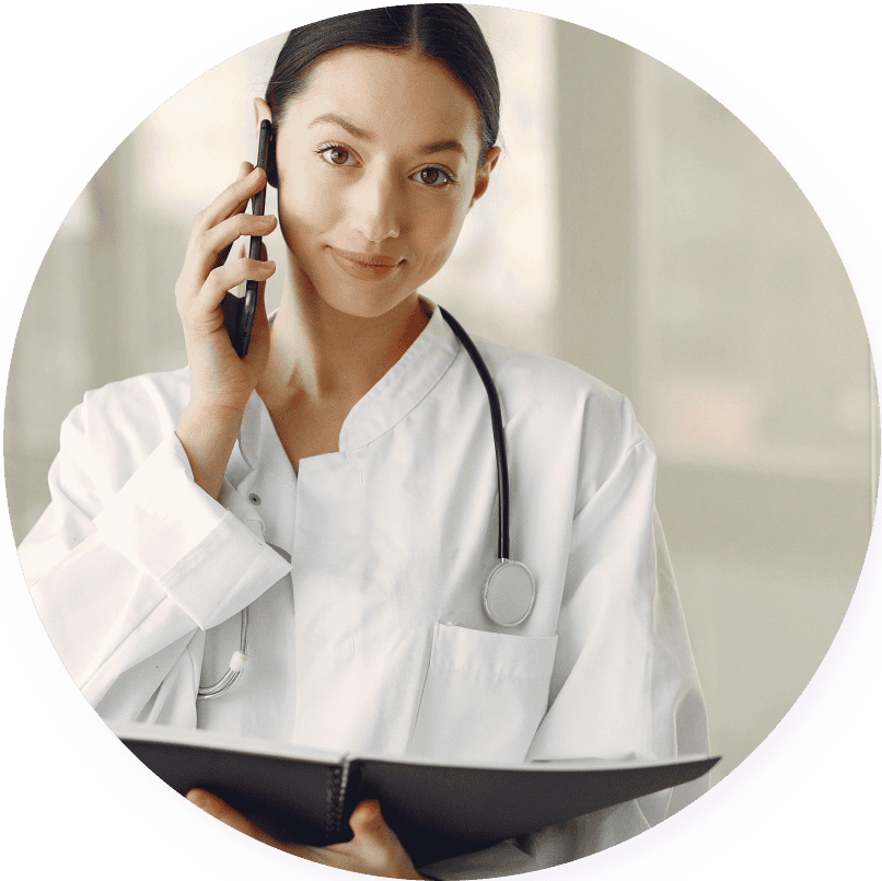 A primary care doctor in a white coat holds a phone to her ear while looking at a file, with a stethoscope around her neck.