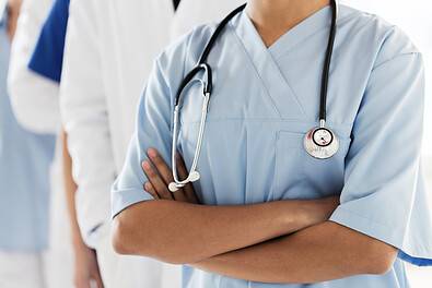A primary care professional in blue scrubs and a stethoscope stands with arms crossed, alongside other medical workers in white coats in the background.
