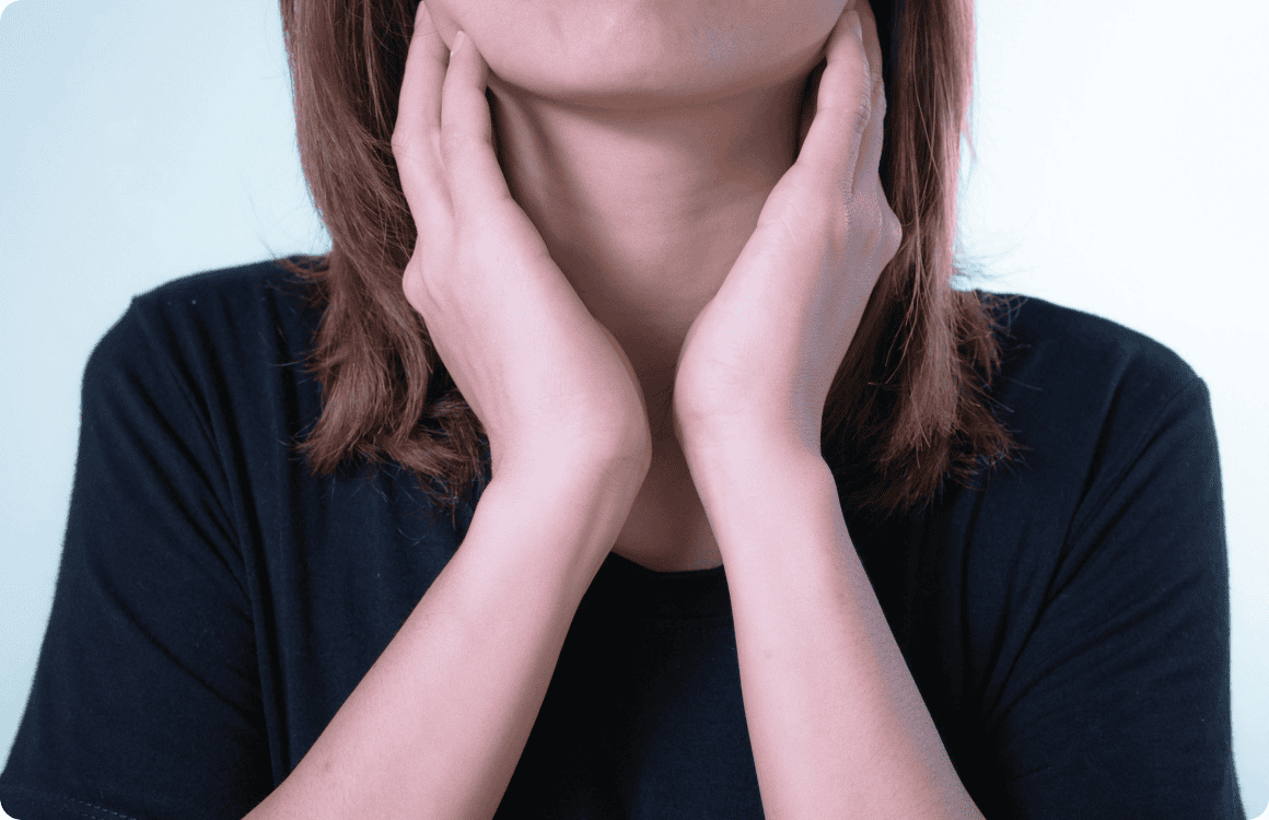 A person with shoulder-length hair, wearing a black shirt, places both hands on their neck, with palms touching the sides of the throat as though in need of primary care.