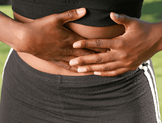 A person wearing black athletic wear holds their hands over their stomach, possibly indicating discomfort or pain, reminding us of the importance of seeking primary care for such symptoms.